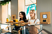 Side view of young cheerful best friends receiving trays with delicious food and beverages in cafeteria