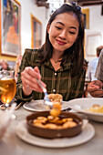 Zufriedene junge ethnische Frau mit langen dunklen Haaren in legerer Kleidung, die lächelnd am Tisch im Restaurant sitzt und leckere Garnelen isst