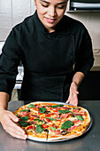 Young positive chef woman in black uniform of tasty fresh cooked pizza placed on table