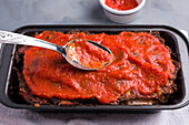 From above of spoon with delicious tomato sauce covering meat in black baking pan placed on gray table
