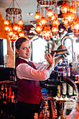 Seitenansicht einer Barkeeperin in Uniform, die einen Cocktail in einem Metallshaker schüttelt, während sie am Tresen eines Pubs steht und in die Kamera schaut