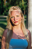 Glückliche Frau mit blondem Haar und Schatten im Gesicht steht an einem sonnigen Tag im Sommerpark und schaut in die Kamera