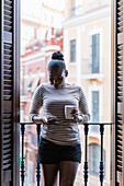 Afroamerikanerin mit einer Tasse Heißgetränk surft im Internet auf einem Handy zwischen Fensterläden zu Hause