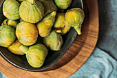 Frische und reife süße grüne Feigen, in einer schwarzen Schale auf dem Holzteller serviert auf dem Tisch mit blauer Tischdecke, Bio-Obst der Saison. Auch bekannt als reife weiße Feigen