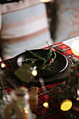 Weihnachtlich gedeckter Tisch mit Kranz auf Keramikteller auf rot kariertem Tischtuch im Hintergrund mit Lichtern