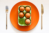 Draufsicht auf eine Kartonschachtel mit Eiern auf einem orangefarbenen Keramikteller auf einem weißen Tisch mit Messer und Gabel