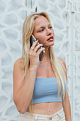 Ruhige Frau mit blondem Haar und in Sommerkleidung steht in der Stadt und spricht mit dem Handy, während sie wegschaut
