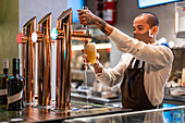 Männlicher Barkeeper in Uniform und Schutzmaske füllt ein Glas mit Bier aus dem Zapfhahn, während er am Tresen in einer Kneipe während einer Coronavirus-Pandemie arbeitet
