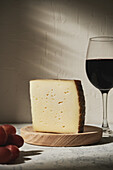 Triangle piece of delicious fresh hard cheese placed on wooden board near grapes and red wine on table in light studio