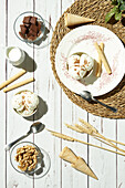 From above of delicious ice cream served in bowls with waffle rolls and cones and placed on white wooden table near pitcher with milk chocolate bars and walnuts