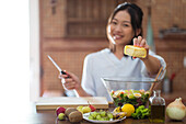Happy Asian female demonstrating piece of cheese over bowl with healthy salad while cooking lunch in sunlit kitchen