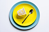 Draufsicht auf einen Joghurtbecher und einen silbernen Löffel auf gelben und blauen Keramiktellern auf einem weißen Tisch in der Küche