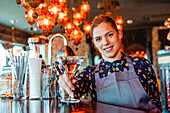 Cheerful female barkeeper with glass of refreshing vermouth standing at counter in bar and looking at camera