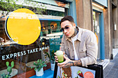 Cool male in sunglasses with healthy drink to go looking away while leaning on urban signboard with photo of food
