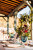 Nahaufnahme einer gedeckten Festtafel mit Kristallgläsern, Besteck und Serviette auf einem Teller neben einem Strauß frischer Blumen für eine Hochzeit und einer Menükarte