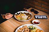 Frau in Freizeitkleidung mit Smartphone und Sonnenbrille isst leckeres taiwanesisches Bento mit Stäbchen in einem asiatischen Cafe
