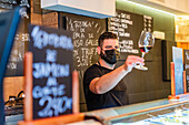 Unscharfer Barmann mit steriler Maske steht an der Theke und schaut in ein Weinglas, während er in einem Restaurant arbeitet