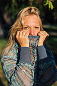 Delicate female with blond hair and in knitted sweater looking at camera in park at sundown