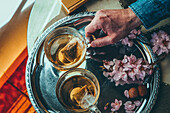 From above hand of crop anonymous man taking glass cup of tea placed on metal tray with flowers in light room