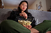 Young Asian female wearing casual clothes sitting with crossed legs on couch and eating popcorn at home