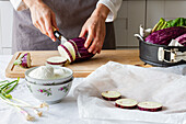 Unbekannter Koch in Schürze schneidet Aubergine mit Messer auf Schneidebrett, während er gesundes Mittagessen in der Küche zubereitet