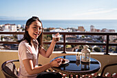 Glückliche Asiatin mit einem Glas Heißgetränk in der Hand, die in die Kamera schaut, während sie beim Frühstück auf dem Balkon am Tisch sitzt