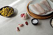 Frische Knoblauchzehen und Salz auf dem Tisch neben einem Seehechtfilet und einer Schüssel mit Erbsen bei der Zubereitung von Speisen in der Küche (von oben)
