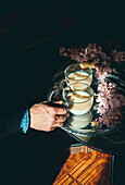 From above hand of crop anonymous person taking glass cup of tea with milk served on tray with flowers in room with sunbeam