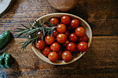 Top view of bowl with fresh cherry tomatoes near rosemary stems and whole onion on wooden table