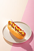 Von oben ein appetitlicher Hot Dog in einem runden Teller auf weißem und rosa Hintergrund