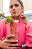 Verträumte Frau in rosafarbenem Pullover sitzt an einem Tisch mit einem köstlichen kalten Beerencocktail in einer Bar im Freien und schaut weg