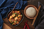 Close up mala tofu, chinese vegan dish, accompanied by a bowl of rice on top of a wooden table decorated with fabrics