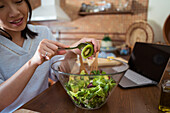Glad Asian woman in casual clothes smiling and putting kiwi into glass bowl with lettuce and avocado while cooking vegan salad for lunch in sunlit kitchen