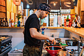 Seitenansicht eines Mannes mit Kopftuch, der an der Theke steht und Ramen in einem modernen asiatischen Café kocht