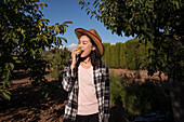 Delighted ethnic female farmer in straw hat and checkered shirt eating fresh tasty apple while standing in orchard in countryside on sunny day