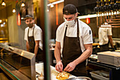Male cook wearing uniform and mask for coronavirus prevention adding cheese on baked flatbread while working in restaurant