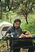 Female mycologist sitting at table against camping tent and taking mushrooms out of wicker basket in woods
