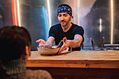 Bärtiger Mann mit Kopftuch und schwarzem T-Shirt stellt einen Teller mit Nudeln auf den Holztresen für einen Kunden in einer Ramen-Bar