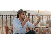 Modern male in stylish outfit and sunglasses sitting on cushions and taking picture of glass bottle of beer on cellphone on terrace bar in Cappadocia, Turkey
