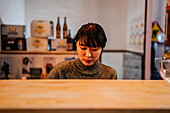 Junge asiatische Frau in Freizeitkleidung sitzt am Holztresen und wartet auf ihre Bestellung in einer Ramen-Bar