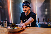 Bearded male in bandana and black t shirt putting plate with noodle on wooden counter for client in ramen bar