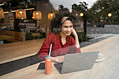 Blick durch eine Glasscheibe auf eine junge Bloggerin, die in einem modernen Restaurant an einem Tisch mit einer gesunden Mahlzeit sitzt und einen Laptop benutzt