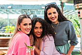 Smiling young multiracial female friends in casual clothes embracing and looking at camera while spending sunny day together on outdoor restaurant terrace