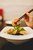 Hand einer Frau, die am Tisch sitzt und mit Stäbchen chinesische Fleischrollen von einem weißen Keramikteller isst