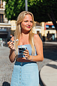 Beautiful blonde young female eating cold tasty ice cream while standing in city street on sunny day in summer