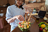 High angle of crop Asian female putting fresh shrimps on top of salad in glass bowl while following recipe from internet in kitchen at home