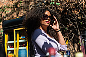 Side view gorgeous young ethnic female touching stylish sunglasses and looking at camera while standing in sunny backyard