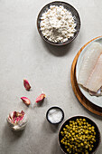 Frische Knoblauchzehen und Salz auf dem Tisch neben Seehechtfilet und einer Schüssel mit Erbsen bei der Essenszubereitung in der Küche von oben gesehen