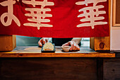 Bildausschnitt Person, die ein asiatisches Gericht mit Soße auf ein Holzbrett mit rotem Tuch am Fenster eines Cafés stellt