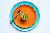 From above of half of fresh ripe juicy kiwi and spoon placed on orange and blue plates on white surface
